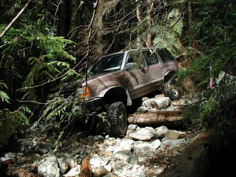 1985 Ford Bronco - A brown SUV with off-road tires navigates a rocky, wooded trail, with its front tires climbing over logs and rocks.