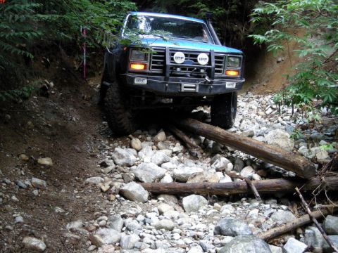 1985 Ford Bronco - A blue 1980s Ford Bronco is navigating a rocky, wooded trail, with logs blocking its path. The off-road vehicle has aftermarket IPF lights…