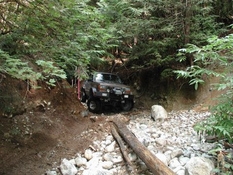 1985 Ford Bronco - A lifted brown 1985 Ford Bronco is navigating a rocky, muddy trail in a wooded area. The vehicle is equipped with off-road tires and a winc…