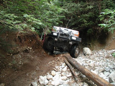 1985 Ford Bronco - A black Ford Bronco is navigating a muddy and rocky trail in a forest setting, with its front right tire climbing over a fallen log.