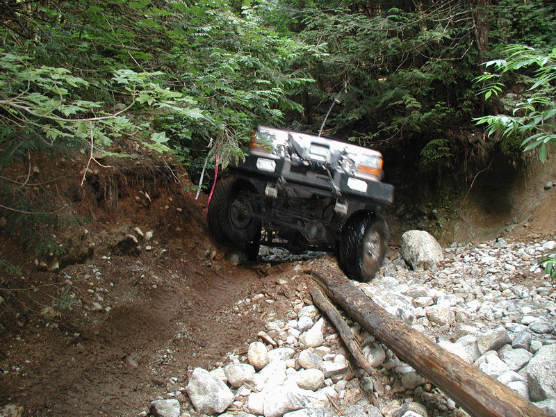 Black Ford Bronco off-roading on a muddy, rocky forest trail, climbing over a log.