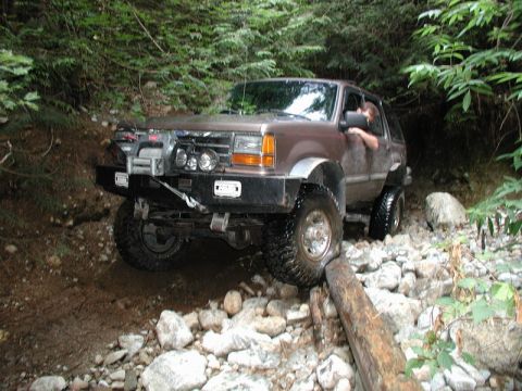 1985 Ford Bronco - A brown SUV with a winch and aftermarket bumper navigates a rocky, wooded trail, with its front tire resting on a fallen log.