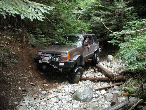 1985 Ford Bronco - A brown SUV with a winch and off-road tires navigates a rocky, wooded trail with a driver looking out the window.