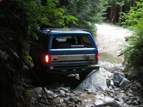 1985 Ford Bronco - The rear of a blue Ford Bronco is shown navigating a rocky, wooded trail with a small stream crossing. The vehicle is positioned on large r…