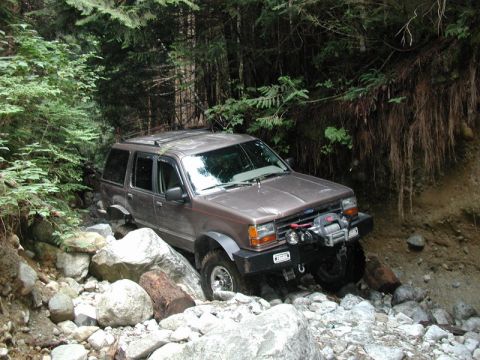 1985 Ford Bronco - A brown SUV is navigating a rocky, wooded trail with a winch mounted on the front bumper.