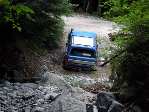 1985 Ford Bronco - A blue Ford Bronco is driving through a shallow stream on a dirt road surrounded by trees. The vehicle has a snorkel and luggage in the bac…