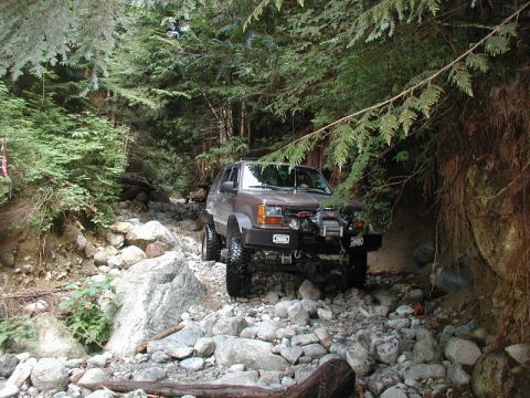 1985 Ford Bronco - A modified brown SUV navigates a rocky, wooded trail. The vehicle features large off-road tires, a winch, and auxiliary lights on the front…