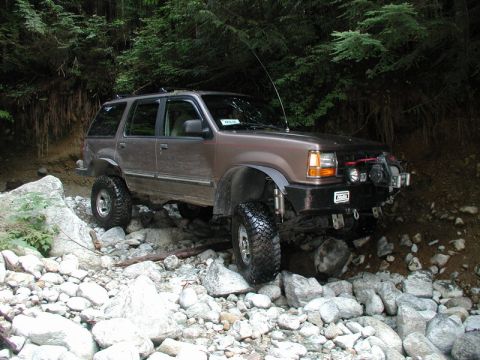1985 Ford Bronco - A lifted, brown SUV is navigating a rocky, wooded terrain. The vehicle has large off-road tires and a custom front bumper with a winch and…