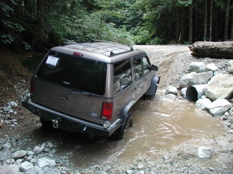 1985 Ford Bronco - A tan Ford Explorer is driving through a shallow, muddy stream on a gravel road surrounded by trees. The vehicle is angled towards the righ…