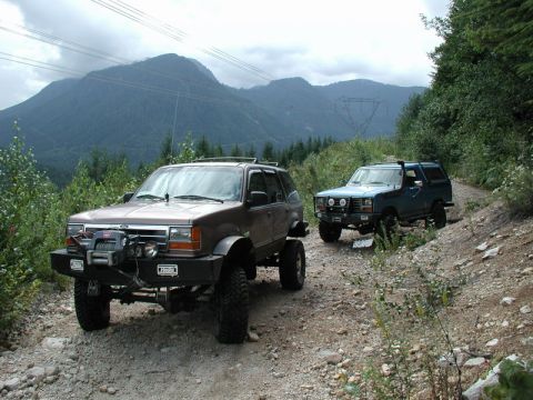 1985 Ford Bronco - Two modified off-road vehicles, a brown SUV in the foreground and a blue SUV behind it, drive up a gravel road in a mountainous, forested a…