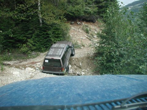 1985 Ford Bronco - A brown SUV navigates a steep, rocky off-road trail with dense green trees on either side. The view is from the perspective of another vehi…