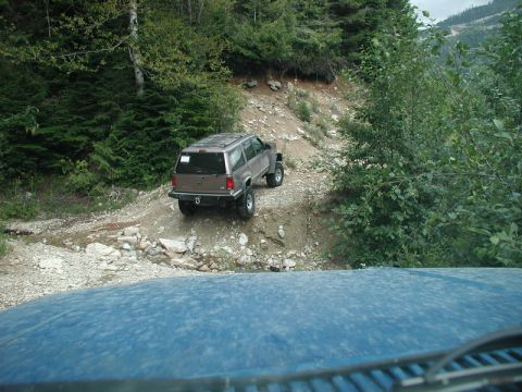 1985 Ford Bronco - A brown SUV is driving up a rocky, wooded trail. The photo is taken from the perspective of another vehicle driving behind it.