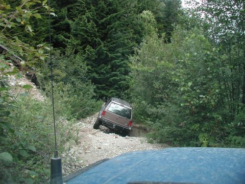 1985 Ford Bronco - A brown SUV navigates a steep, rocky, off-road trail surrounded by dense forest. The front of another vehicle, likely the one taking the ph…