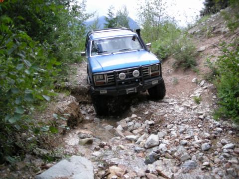 1985 Ford Bronco - A blue 1985 Ford Bronco is navigating a rocky, off-road trail with water flowing over the path. The vehicle is equipped with a front brush…