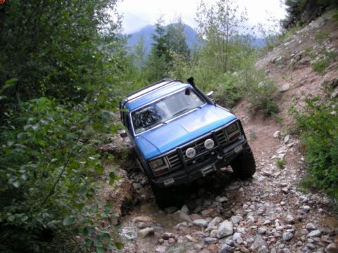 1985 Ford Bronco - A blue 1980s Ford Bronco is navigating a rocky, off-road trail with dense foliage on the left and a steep embankment on the right. The vehi…
