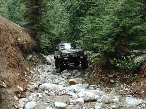 1985 Ford Bronco - A dark-colored SUV navigates a rocky, off-road trail surrounded by dense forest. The vehicle appears to be modified for off-roading with la…