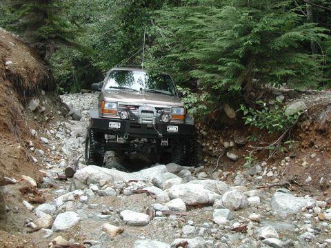 1985 Ford Bronco - A tan 1980s Ford Bronco is navigating a rocky creek bed surrounded by trees. The off-road vehicle features a winch and aftermarket bumper.