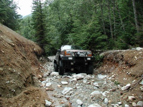 1985 Ford Bronco - A brown 1980s SUV drives through a rocky stream bed in a forest. The vehicle has a winch and brush guard on the front bumper.