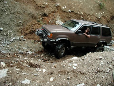 1985 Ford Bronco - A brown SUV with large off-road tires navigates a rocky, muddy terrain, with a driver visible through the open window. The vehicle appears…