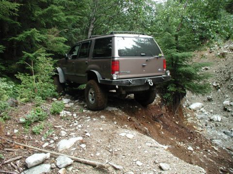 1985 Ford Bronco - A lifted, brown Ford Explorer is navigating a steep, rocky, and wooded trail. The vehicle is angled on the incline, with its rear facing th…