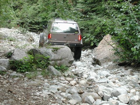 1985 Ford Bronco - A tan Ford Explorer SUV navigates a rocky, wooded trail, with large boulders and dense foliage surrounding the path.