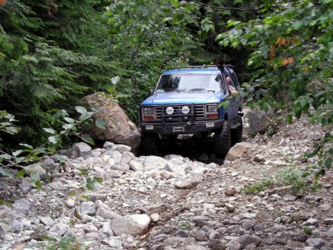 1985 Ford Bronco - A blue 1980s Ford Bronco navigates a rocky, wooded trail. The off-road vehicle is equipped with auxiliary lights and a brush guard.
