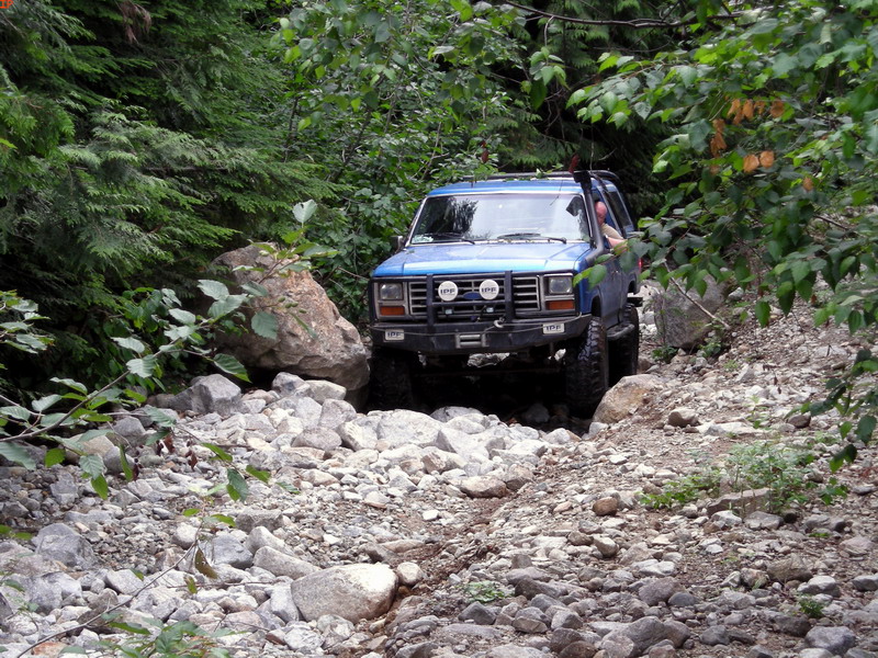 Blue 1980s Ford Bronco off-roading on a rocky trail surrounded by trees.
