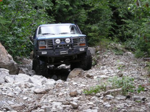 1985 Ford Bronco - A blue 1980s Ford Bronco is navigating a rocky, wooded trail. The vehicle has a winch and auxiliary lights mounted on the front bumper.