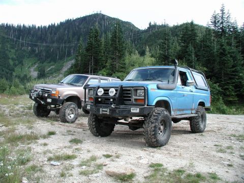 1985 Ford Bronco - Two modified off-road SUVs are parked on a gravelly surface with a forested mountainside in the background. The blue SUV in the foreground…