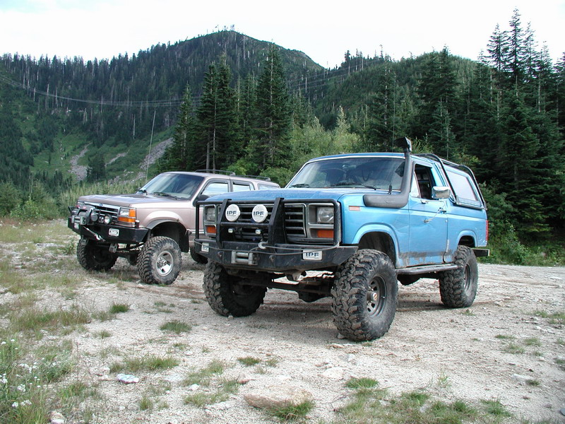 Two modified off-road SUVs parked on gravel, with a forested mountain in the background.