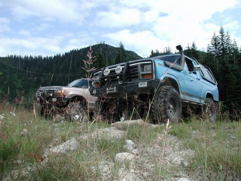1985 Ford Bronco - Two modified off-road SUVs are parked on a rocky, grassy hillside with a forest and power lines in the background. The SUV in the foregroun…