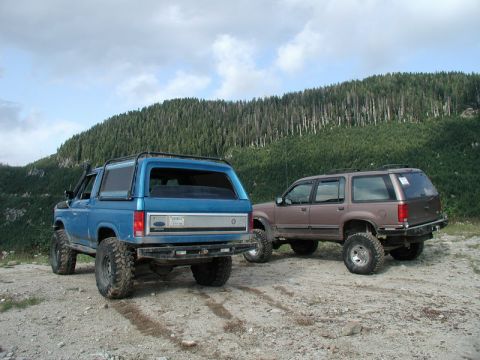 1985 Ford Bronco - A lifted blue 1980s Ford Bronco and a lifted brown Ford Explorer are parked on a dirt road with a forested mountain in the background.