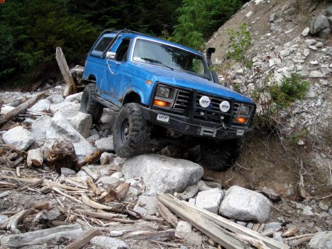 1985 Ford Bronco - A blue Ford Bronco is navigating a rocky, off-road trail with large boulders and fallen trees. The vehicle has large off-road tires and aux…