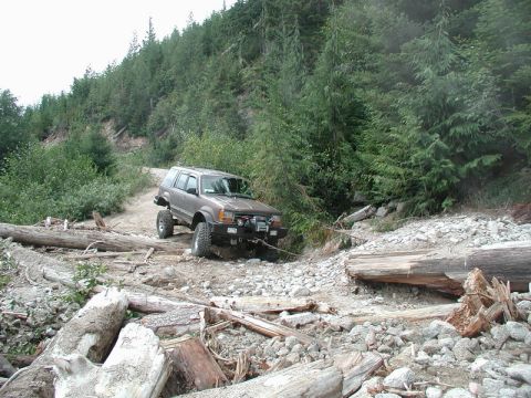 1985 Ford Bronco - A lifted, brown SUV navigates a rough, rocky, and wooded terrain with fallen logs obstructing the path. The vehicle appears to be winched t…