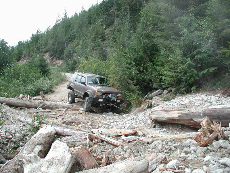 Lifted brown SUV on a rocky, wooded trail with fallen logs.