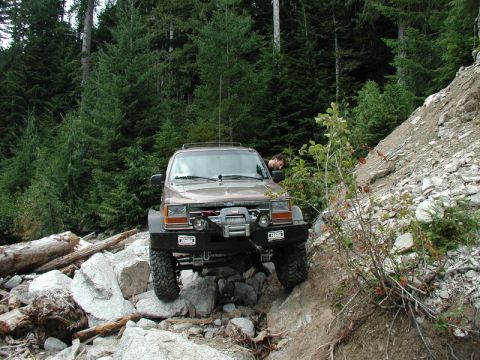 1985 Ford Bronco - A brown Ford Explorer is navigating a rocky, off-road trail with a dense forest in the background. The vehicle features a winch and afterma…