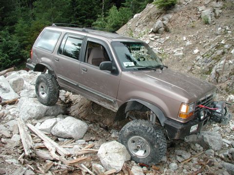 1985 Ford Bronco - A lifted, brown Ford Explorer is navigating a rocky, off-road trail with its front tire on a large boulder and its rear tire on a rock. The…