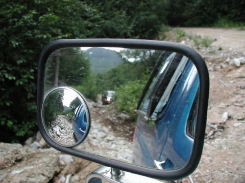 1985 Ford Bronco - A blue Ford Bronco is seen from the side mirror, driving on a rocky, unpaved road through a forest. The side mirror also shows a smaller co…