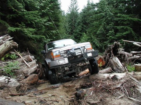 1985 Ford Bronco - A brown 1980s SUV with a winch is navigating a muddy, debris-strewn forest trail, with large fallen logs and dense evergreen trees surround…