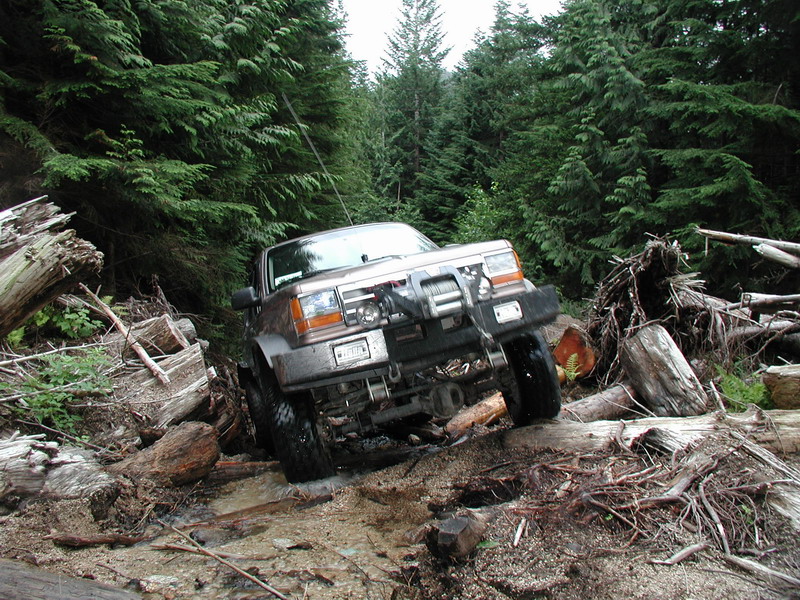 Brown 1980s SUV with winch on a muddy forest trail with logs and trees.
