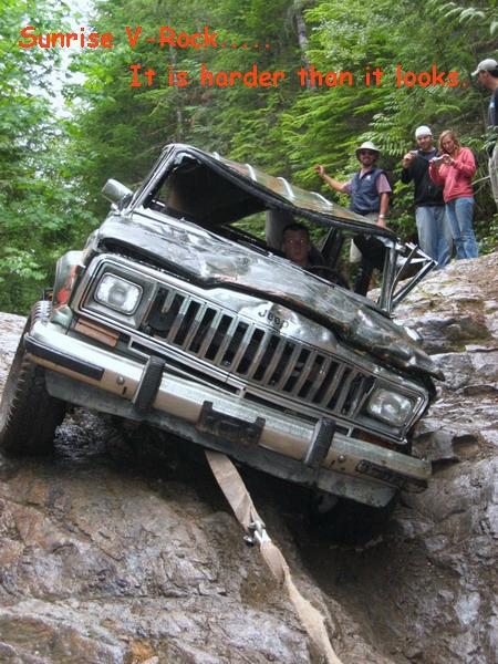 1985 Ford Bronco - A heavily damaged Jeep Cherokee is stuck on a steep, rocky incline, with a tow strap attached to its front bumper. Several people are stand…