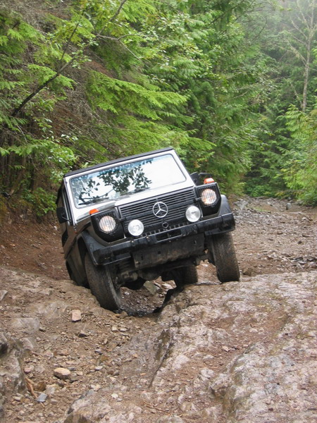 1985 Ford Bronco - A black Mercedes-Benz G-Wagen is navigating a steep, rocky off-road trail with its front passenger side wheel significantly elevated. The v…