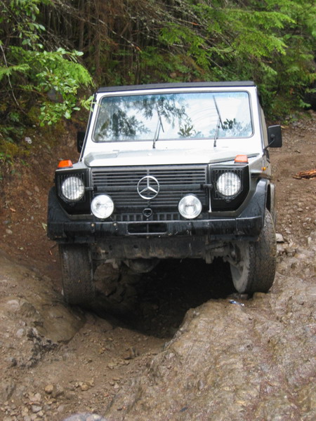 1985 Ford Bronco - A silver Mercedes-Benz G-Wagen is navigating a challenging off-road trail with a deep rut in the foreground.
