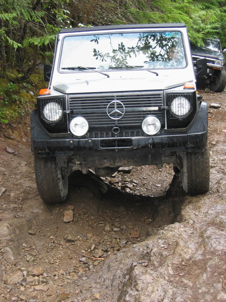 1985 Ford Bronco - A silver Mercedes-Benz G-Wagen navigates a deep rut on a muddy, rocky off-road trail. The front of the vehicle is angled towards the camera…
