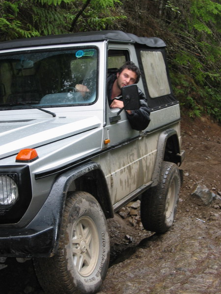 1985 Ford Bronco - A man leans out the driver's side window of a silver off-road vehicle, holding a side mirror. The vehicle is on a dirt track with trees in…