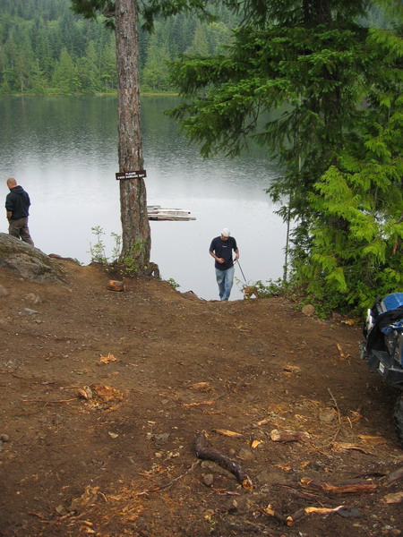 1985 Ford Bronco - A blue ATV is parked on a dirt path next to a lake. A man is walking his dog down towards the water.