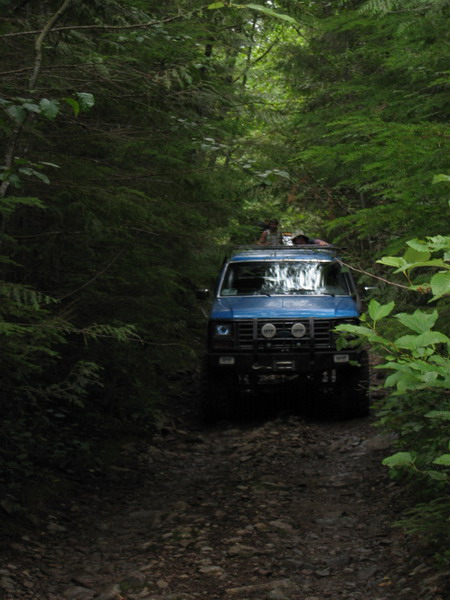 1985 Ford Bronco - A blue SUV drives down a rocky, tree-lined dirt path in a forest. Several people are visible in the open sunroof of the vehicle.