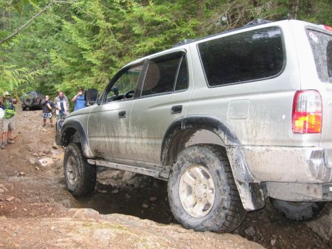 1985 Ford Bronco - A silver SUV navigates a rocky, muddy trail in a forest setting. Several people are standing around the vehicle and in the background.