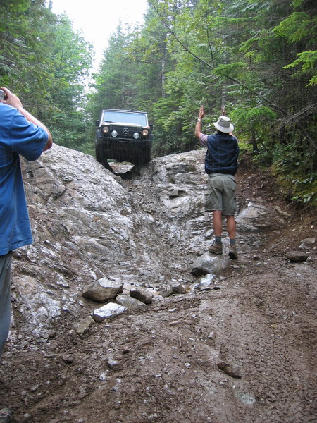 1985 Ford Bronco - A black SUV navigates a steep, rocky, and muddy incline in a forest. A person in a hat stands on the side of the trail, gesturing.