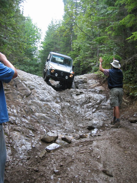 1985 Ford Bronco - A silver Mercedes-Benz G-Wagen is navigating a steep, rocky, and muddy off-road trail in a forest. A person in a hat and vest is guiding th…